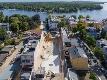 View of the elongated, almost completed excavation pit from above. Immediately to the left and right of the excavation pit are 3-storey apartment blocks. Several pieces of construction equipment are still in use. A short side of the excavation pit adjoins the road. After another row of houses is a wide river. On the other side of the river is a deciduous forest. On the horizon is a hill with a wind turbine.