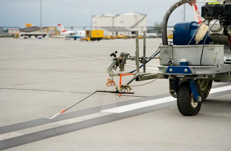Photo: Road marking: A vehicle equipped with a spraying device applies white colour to a masked area of an airport runway.